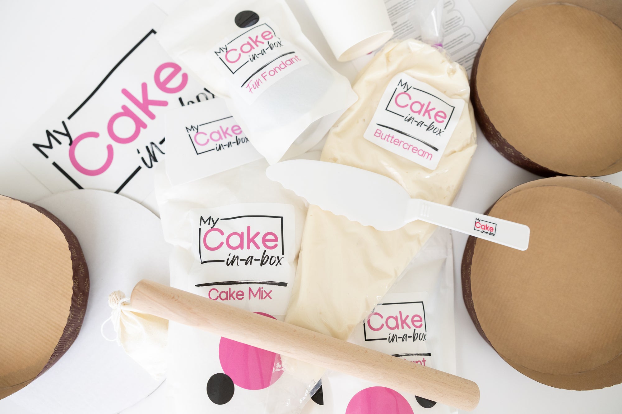 A styled flat-lay of a cake mix kit by My Cake in-a-Box, featuring multiple white bags of cake mix, buttercream, and cake mix with pink and black branding, a white spatula, a wooden rolling pin, and round brown cardboard cake bases on a white background.