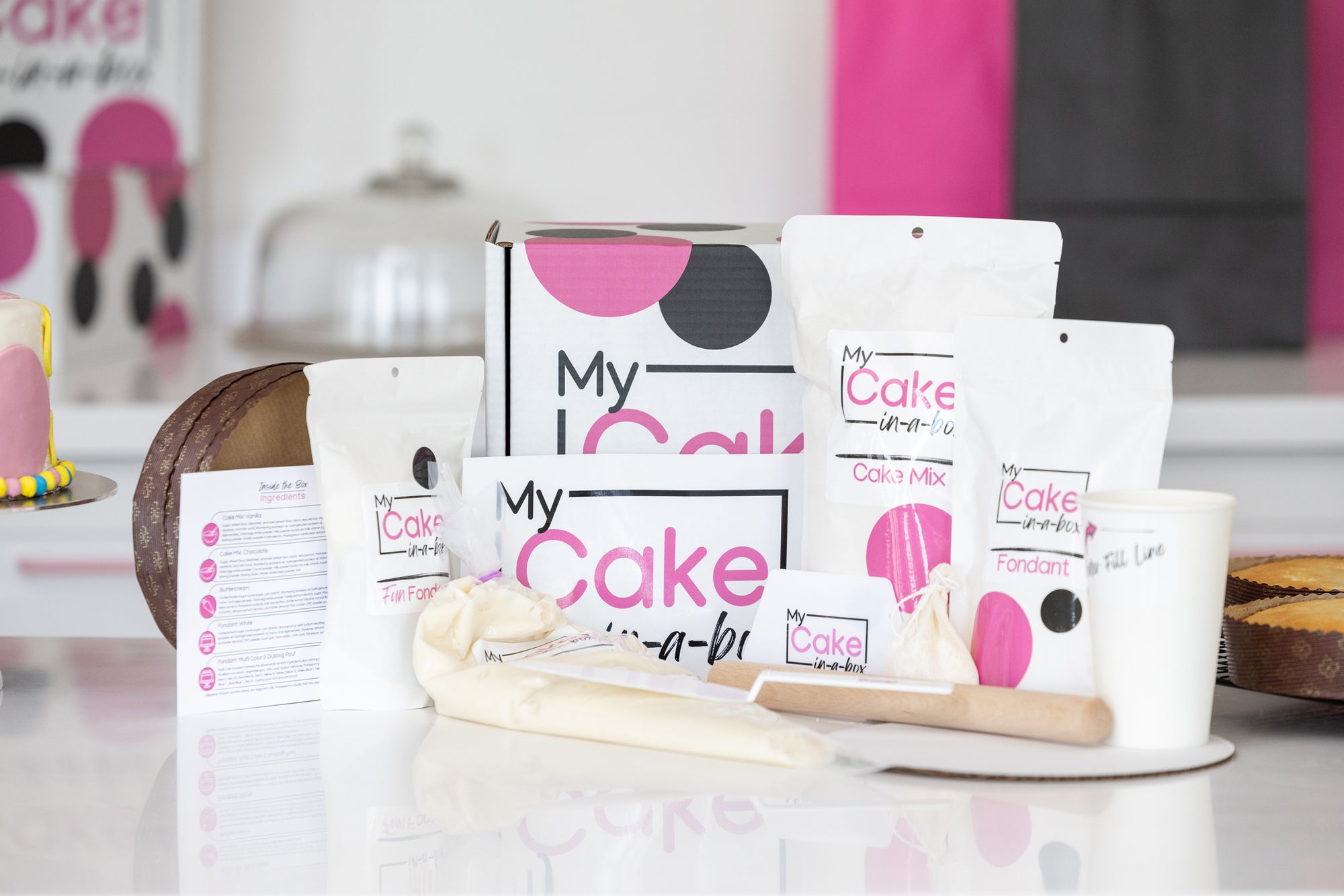 A baking & decorating kit set on a white reflective surface, featuring 'My Cake-in-a-Box' labeled white pouches of cake mix, fondant, and butter, a box, a piping bag, a wooden spatula, and a small white cup, with a blurred kitchen background.