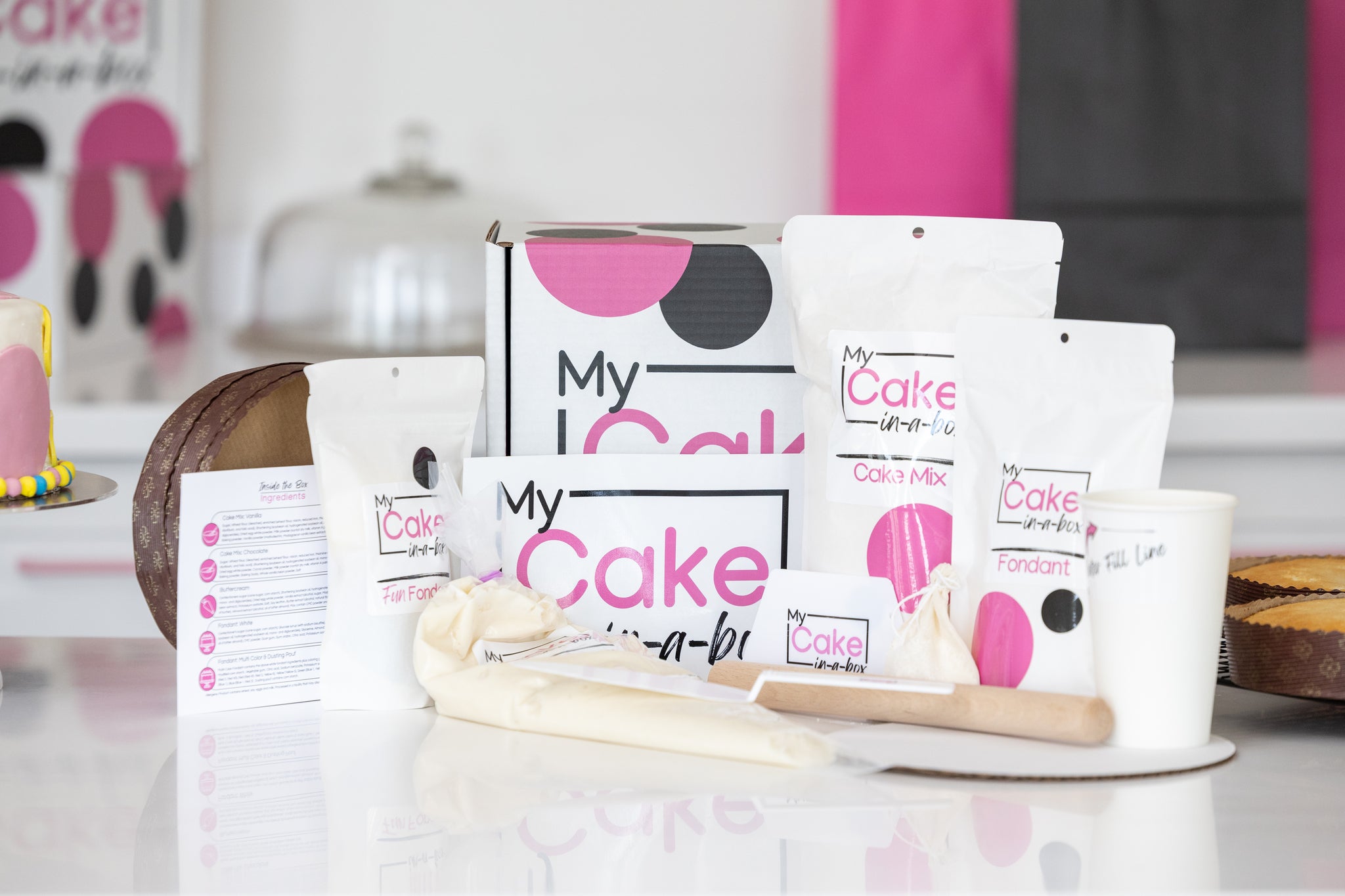 A baking & decorating kit set on a white reflective surface, featuring 'My Cake-in-a-Box' labeled white pouches of cake mix, fondant, and butter, a box, a piping bag, a wooden spatula, and a small white cup, with a blurred kitchen background.