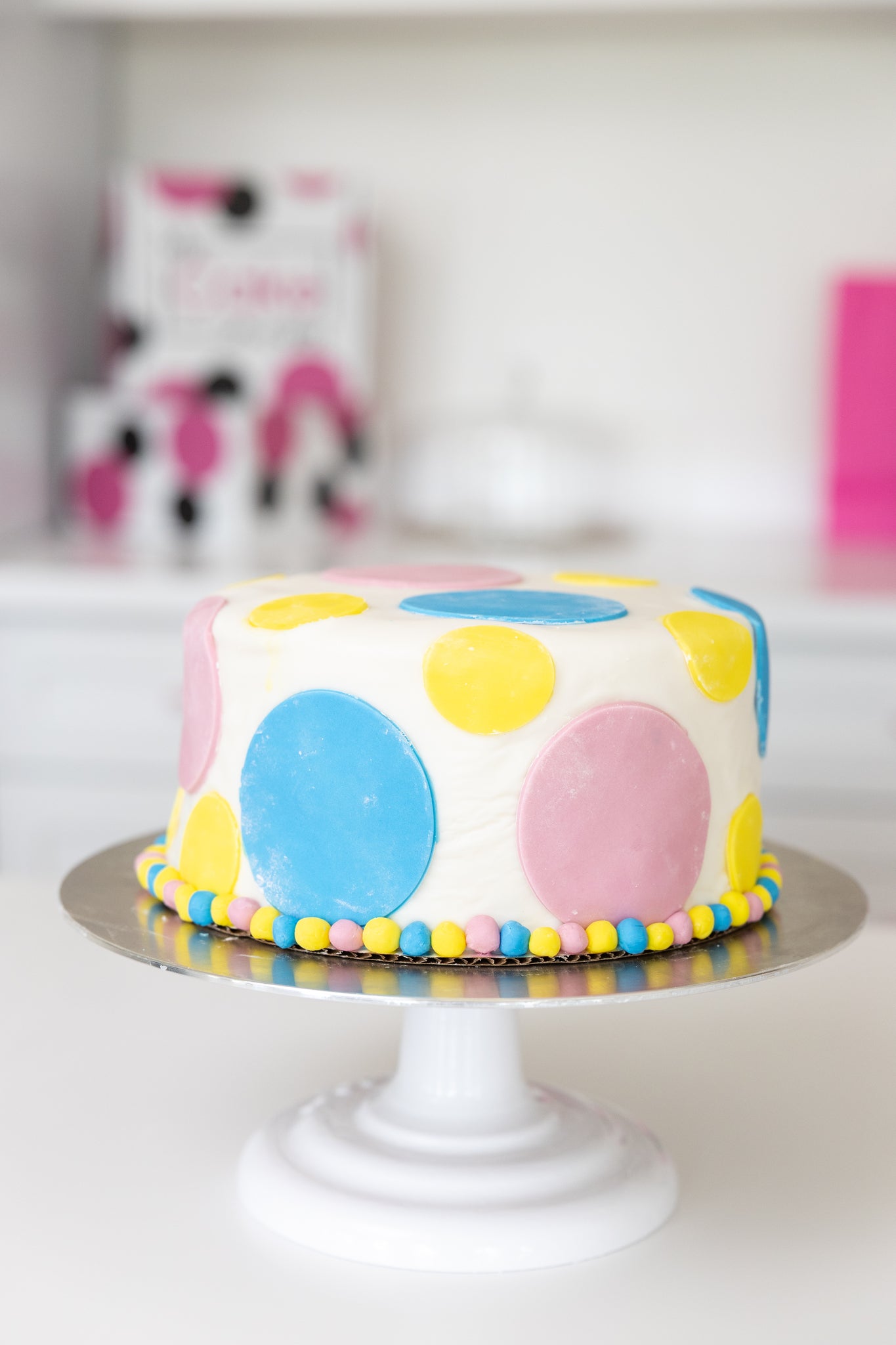 A round white fondant covered cake with large pastel pink, blue, and yellow polka dots, sitting on a white pedestal cake stand with a silver and white base.